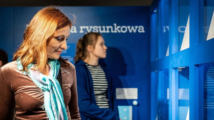 Dwie kobiety. Na pierwszym planie kobieta w średnim wieku, rudowłosa patrzy się przez siebie, podziwiając muzealny obiekt. Na drugim planie młoda kobieta również coś ogląda, patrząc przed siebie. Ściany pomieszczenia są niebieskie. 